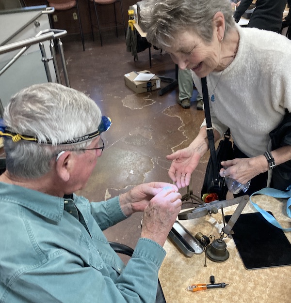 A man with short great hair helps a woman with short tan hair inspect a broken device